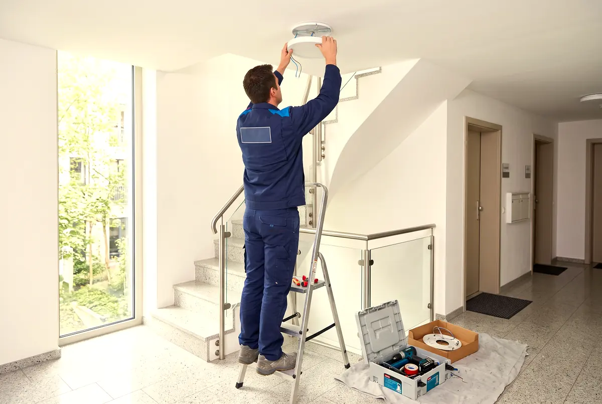 Hausmeister auf einer Stehleiter montiert eine neue Deckenleuchte im hellen Treppenhaus einer Wohnanlage